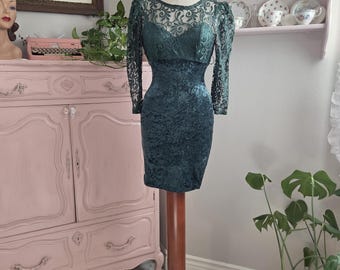 May include: A dark teal, vintage-style dress on a wooden mannequin. The dress features a velvet-like texture on the bodice and skirt, with lace sleeves and neckline. The dress is knee-length and has a fitted silhouette.