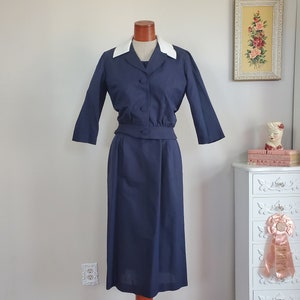 May include: A navy blue dress with a white collar and a fitted bodice. The dress has a skirt that falls to the knee and is made of a smooth fabric.