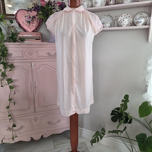 May include: A vintage, pale pink nightgown displayed on a wooden dress form. The nightgown features a Peter Pan collar, short sleeves with lace detailing, and a button-down front. The background includes a pink cabinet, floral arrangements, and decorative plates.