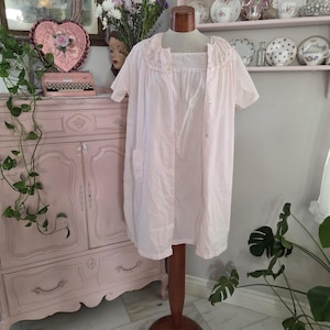 May include: A light pink vintage nightgown with short sleeves and a button-down front. The nightgown has a square neckline and a ruffled collar with pink embroidery. It is displayed on a wooden mannequin stand in a room with vintage decor.