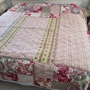 May include: A patchwork quilt featuring a variety of fabric patterns including floral prints in shades of red, pink, and green, alongside striped and textured sections. The quilt is stitched with a grid pattern and covers the surface of a bed.
