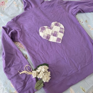 Puede incluir: Una sudadera con capucha morada con un corazón a cuadros blanco y morado aplicado en la espalda. La sudadera tiene una capucha con cordón y mangas largas. Un pequeño ramo de flores blancas y hojas verdes descansa sobre la manga.
