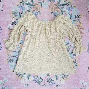 May include: A cream-colored lace top with a ruffled neckline and 3/4 sleeves. The top is made of a delicate lace fabric and has a relaxed fit.