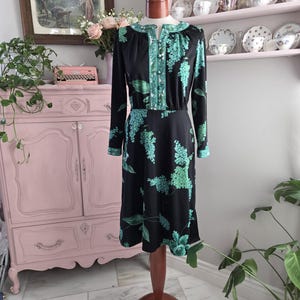 May include: A vintage black dress with a floral pattern in shades of green and teal. The dress has long sleeves, a button-up front, and a round neckline with teal trim. The dress is displayed on a wooden dress form.