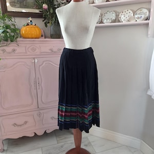 May include: A black pleated skirt with a colourful striped hem is displayed on a mannequin. The stripes include red, green, and blue. The mannequin is on a wooden stand in a room with a pink cabinet and decorative plates.