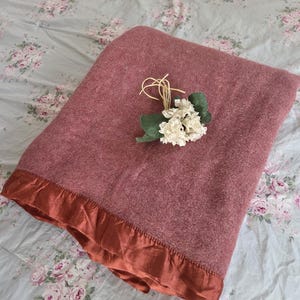 Op de afbeelding: Een stoffig roze deken met een roestkleurige satijnen rand. De deken is opgevouwen en versierd met een klein bosje witte bloemen en touw. De deken ligt op een bloemig patroon beddensprei.