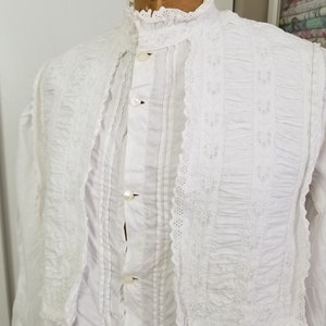 May include: White cotton shirt with a high collar and lace trim. The shirt has a button-up front and is pleated in the front. The lace trim is embroidered with a floral pattern.