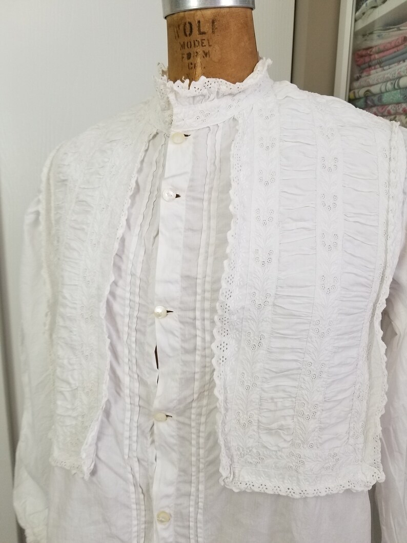 May include: White cotton shirt with a high collar and lace trim. The shirt has a button-up front and is pleated in the front. The lace trim is embroidered with a floral pattern.