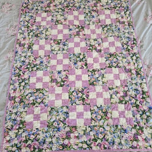 May include: A floral quilt with a checkerboard pattern in lavender and white. The quilt features a border and backing of colorful flowers in shades of pink, purple, and blue. The quilt is a square shape.