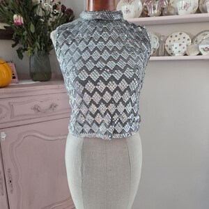 May include: A silver sequined top with a high collar, displayed on a mannequin. The top features a geometric pattern of shimmering sequins. The mannequin is set against a backdrop of vintage decor, including a pink cabinet and shelves of teacups.