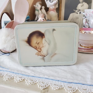 Puede incluir: Una caja de hojalata vintage con un diseño azul y blanco. La tapa presenta una pintura de un bebé durmiendo sosteniendo un animal de peluche blanco.