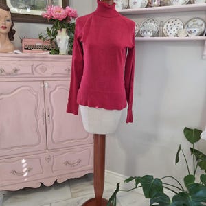 以下が含まれることがあります： マネキンに展示された赤いタートルネックセーター。長袖セーターはリブ編みの裾と袖口が付いています。マネキンは丸いベースの木製スタンドにあります。背景には、ピンクのキャビネット、アートワーク、装飾プレートが含まれています。