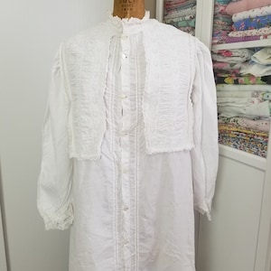 May include: A white cotton nightgown with a lace trim and a button-up front. The nightgown has a high neckline and long sleeves.