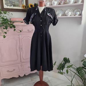 Könnte beinhalten: Ein schwarzes Vintage-Kleid mit Kragen und Knopfleiste vorne. Die kurzen Ärmel und der Kragen haben ein Karomuster. Das Kleid wird auf einer Schaufensterpuppe mit Holzständer präsentiert.