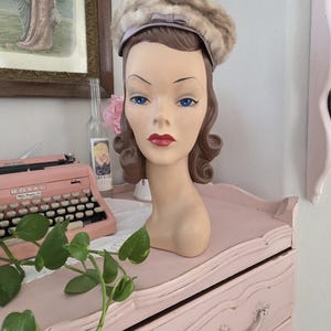 May include: A vintage-style hat on a mannequin head. The hat is a mix of light brown and beige fur with a satin band and bow. The mannequin has blue eyes and red lips. The head is displayed on a pink dresser.