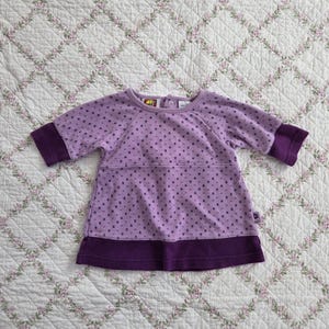 May include: A lavender baby dress with short sleeves and a dark purple hem. The dress features a pattern of small, dark purple hearts and a dark purple band at the sleeves. The dress is made of soft fabric.