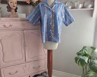 May include: A short-sleeved blue button-down shirt with a floral pattern. The shirt is displayed on a mannequin. The floral pattern includes pink and white flowers with vertical stripes. The shirt has a collar and is buttoned up.