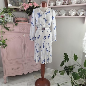 Puede incluir: Un vestido blanco vintage con estampados florales azules y pequeños lunares. El vestido presenta mangas largas, una parte delantera abotonada y una falda plisada. Un cinturón marrón ciñe la cintura. El vestido se exhibe en un maniquí de madera.