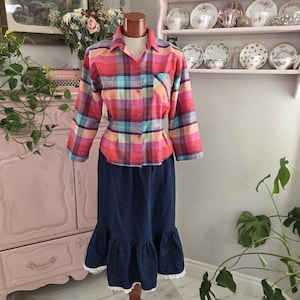 May include: A vibrant plaid shirt with a button-down front and long sleeves, combined with a dark blue denim skirt. The skirt features a ruffled hem with white lace edging. The outfit is presented on a mannequin.