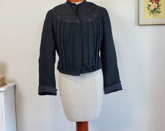 May include: A black cropped jacket with a high collar and pleated front. The jacket has a decorative trim around the collar and buttons.