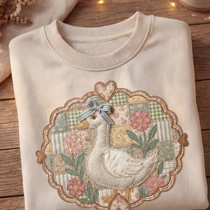 Peut inclure: Sweat-shirt crème avec un motif brodé d'une oie blanche avec un nœud bleu, entourée d'un motif floral patchwork. Le motif comprend des fleurs roses, des feuilles vertes et un cœur. Le sweat-shirt est plié sur une surface en bois.