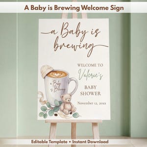 Puede incluir: Un letrero de bienvenida rectangular con el texto A Baby is brewing, Welcome to Valerie's Baby Shower y November 12, 20xx. El diseño incluye una taza de café con arte latte, un pequeño oso de peluche, hojas verdes y una cinta beige.