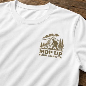以下が含まれることがあります： 白いクルーネックTシャツ。左胸に茶色のグラフィックがあり、山と木々の前で薪を割る人物が描かれています。「MOP UP BUILDS CHARACTER」のテキストが下にあります。