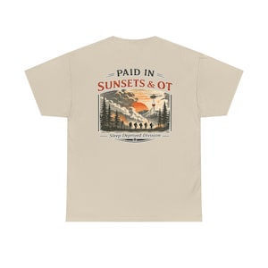 May include: A beige t-shirt featuring a graphic on the back with the text PAID IN SUNSETS & OT and Sleep Deprived Division. The illustration depicts a forest fire scene with silhouettes of people, pine trees, a red sun, and a helicopter.