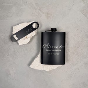 May include: A matte black metal flask engraved with the name Alexander, the word GROOMSMAN, and the date 20.04.2026, paired with a matching black metal bottle opener featuring the engraved initials A.P. in a script font.