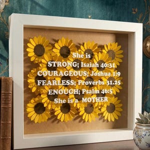 May include: A framed artwork with paper sunflowers. The sunflowers have yellow petals and brown centres. White text reads "She is STRONG; COURAGEOUS; FEARLESS; ENOUGH; She is a MOTHER" with Bible verses. The frame is white.