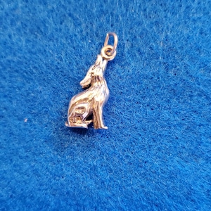 May include: A silver-colored pendant in the shape of a howling wolf. The wolf is depicted in a standing position with its head raised and mouth open. The pendant has a small loop at the top for attaching to a chain.