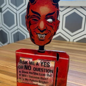May include: A red devil-faced fortune teller with black horns and a winking expression. The base is red and features the text "ASK ME A YES OR NO QUESTION" with three questions: "Does He/She Love Me?", "Will I Become Rich?", and "Is My Future Bright?"