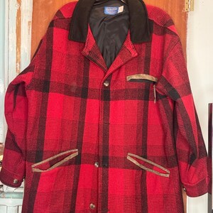 May include: A red and black checked wool jacket with a black collar and lining. The jacket features two front pockets with brown trim and is buttoned. The Pendleton label is visible on the collar.
