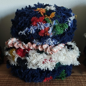 May include: A handmade, textured decorative object. The piece is primarily navy blue, with layers of white, red, and patterned fabric. Accents of yellow, green, and other colors add visual interest. The object is sitting on a wooden surface.