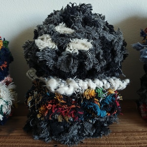 May include: A handmade textile sculpture with a textured, shaggy appearance. The top section is primarily dark gray with white patches, while the base features a vibrant mix of colorful threads, including red, yellow, and blue. The sculpture is displayed on a wooden surface.