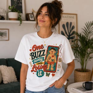 May include: A white t-shirt featuring a colorful graphic print with the text One BUZZ away FROM Losing it. The design includes a cartoon character inside a red frame, surrounded by small bones, hearts, and butterflies in red, blue, and yellow.