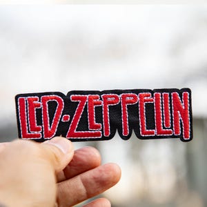 Peut inclure: Un écusson brodé noir avec le texte "LED ZEPPELIN" en rouge, bordé de blanc. L'écusson est rectangulaire avec un bord inférieur ondulé. L'écusson est tenu dans une main.