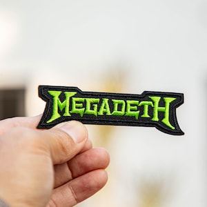 Puede incluir: Un parche bordado negro con la palabra "MEGADETH" en letras verdes brillantes y estilizadas. El parche tiene forma rectangular con extremos puntiagudos. El parche está sostenido en la mano de una persona.