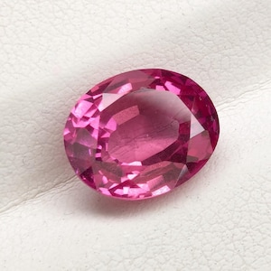 Puede incluir: Una gema rosa facetada, de forma ovalada. La gema está colocada sobre un fondo blanco dentro de una caja negra. Las facetas de la piedra reflejan la luz, creando un efecto brillante. Esta gema es adecuada para la fabricación de joyas.