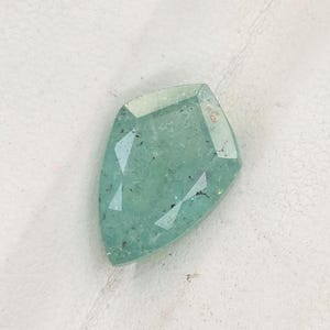 May include: A loose, shield-shaped gemstone with a faceted surface and a pale mint green color. The stone displays internal inclusions and a translucent quality, resting on a white surface within a black-bordered display case.
