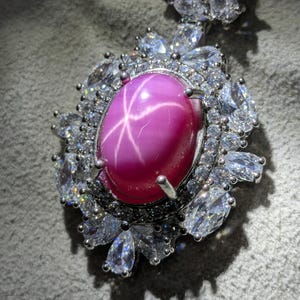 Puede incluir: Colgante de plata con una gran gema rosa ovalada con un patrón en forma de estrella. La gema está rodeada de piedras más pequeñas, claras y brillantes. El colgante tiene un diseño floral.