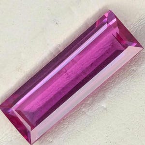 May include: A vibrant, rectangular baguette-cut gemstone in a deep magenta pink hue. The stone features a faceted surface that reflects light, showcasing a clear, transparent quality with sharp, defined edges and a elongated, slender geometric shape.