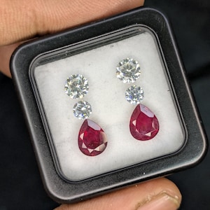 Peut inclure: Une paire de boucles d'oreilles en pierres précieuses présentée dans un étui noir. Chaque boucle d'oreille est ornée d'un rubis rouge foncé en forme de poire, sous deux diamants ronds et clairs. Les boucles d'oreilles sont posées sur un fond blanc.