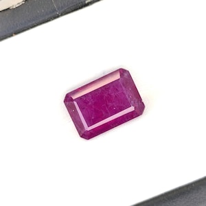 May include: A rectangular, emerald-cut gemstone with a deep, vibrant magenta-pink hue. The stone features faceted surfaces that reflect light, showcasing internal clarity and color saturation, presented against a stark white background.