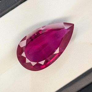 Può includere: Una gemma sfaccettata a forma di pera di un colore rosa magenta intenso e vibrante. La pietra presenta una parte superiore appuntita e una base arrotondata, con molteplici sfaccettature triangolari che riflettono la luce creando brillantezza.