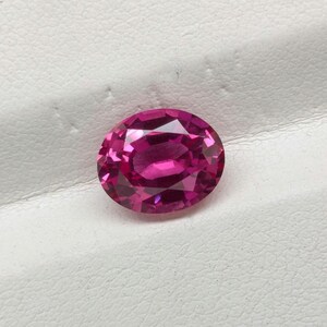 Puede incluir: Una gema de color magenta rosado intenso, con talla ovalada y una superficie facetada que refleja la luz. La piedra muestra una calidad transparente y clara con múltiples facetas geométricas dispuestas en un patrón simétrico.