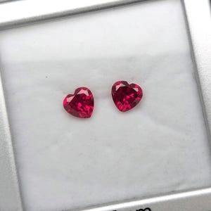 May include: Two vibrant, heart-shaped red gemstones with faceted surfaces, displayed inside a square white presentation box. The box features the printed text 7x7mm, indicating the dimensions of the stones, and has a metallic silver frame.
