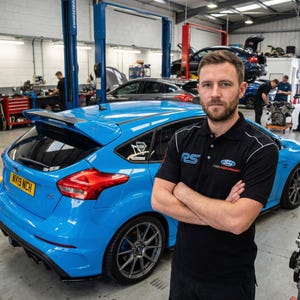May include: A vibrant blue Ford Focus RS hatchback in a garage setting. The car features a rear spoiler and dark alloy wheels. A man wearing a black Ford Performance polo shirt stands in front of the car. Other vehicles and mechanics are visible in the background.