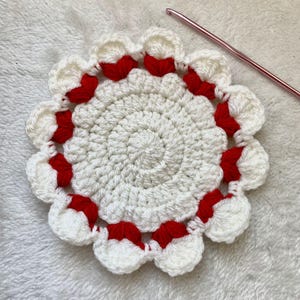 Può includere: Un sottobicchiere all'uncinetto bianco fatto a mano con un bordo smerlato rosso. Il sottobicchiere ha un design circolare con un centro a spirale e un bordo smerlato. Un uncinetto rosa è posto accanto.
