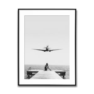 Könnte beinhalten: Schwarz-Weiß-Druck in einem Rahmen, der ein Vintage-Flugzeug zeigt, das über einen Holzsteg fliegt. Eine Person sitzt auf dem Steg und blickt auf das Meer. Das Bild hat eine minimalistische Ästhetik und erzeugt ein Gefühl von Nostalgie.